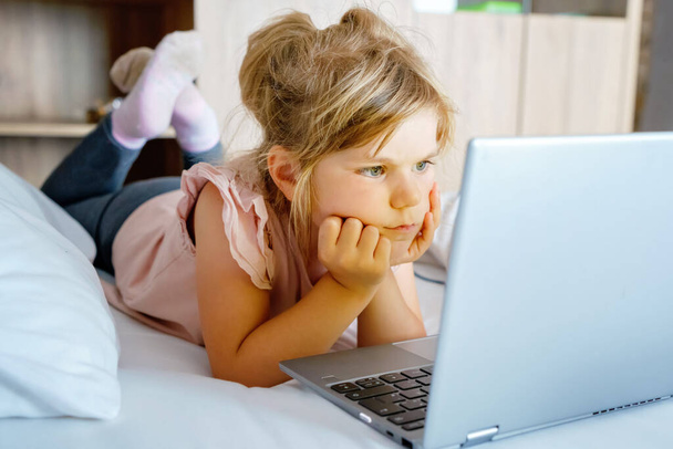 ノートパソコン、ノートブック、ベッドの上できれいな白いリネンのビデオを見て就学前の女の子。子供との屋内活動。フリーランス、遠隔学習、または子供の概念を持つ家庭からの仕事。幸せな子供. - 写真・画像