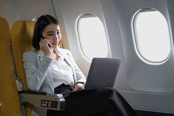 Ασιάτης/ισσα γυναίκα επιβάτης κάθεται σε αεροπλάνο κοντά στο παράθυρο και διαβάζοντας ειδήσεις από τα κοινωνικά δίκτυα ή χρησιμοποιώντας Ταξιδιωτικές εφαρμογές στο smartphone. - Φωτογραφία, εικόνα