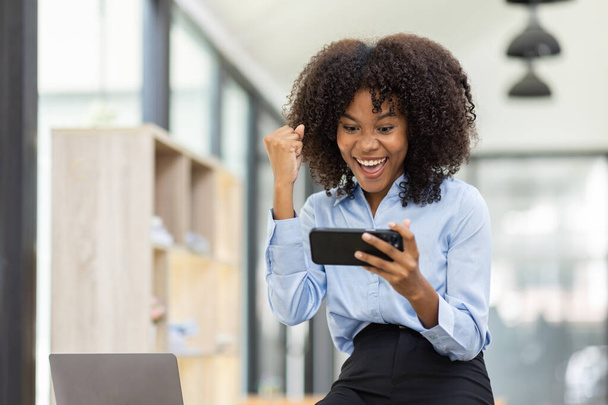 Úspěšná a vzrušující mladá africká americká obchodní žena při pohledu na displej telefonu, africká žena nadšená vyhrát na svém smartphonu v práci. - Fotografie, Obrázek