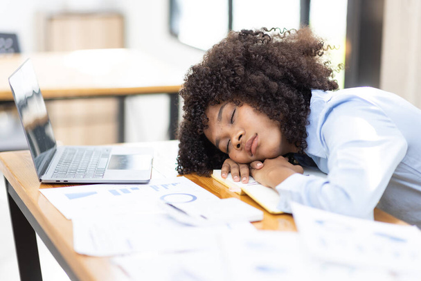 ノートパソコンや書類の横にあるオフィスの机の上で眠っているアフリカの女性は、過労で疲れています。職場での慢性疲労に苦しむ若いアフリカの女の子の仕事中毒 - 写真・画像