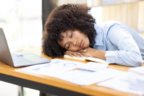 ノートパソコンや書類の横にあるオフィスの机の上で眠っているアフリカの女性は、過労で疲れています。職場での慢性疲労に苦しむ若いアフリカの女の子の仕事中毒 - 写真・画像