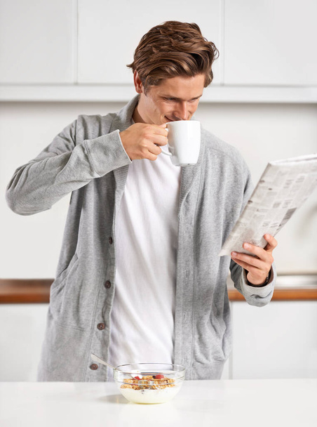 Το πρωινό μου τελετουργικό... ένας όμορφος νεαρός άνδρας πίνει ένα ζεστό ρόφημα και διαβάζει εφημερίδα. - Φωτογραφία, εικόνα