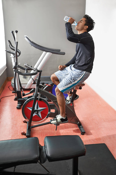 Manter-se hidratado é importante. Um jovem sentado em uma bicicleta de exercício e água potável - Foto, Imagem