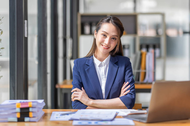 Kahverengi saçlı, açık mavi ceketli, gülümseyen Asyalı kadın ofiste evrak bilgisayarıyla çalışıyor, mali raporu analiz ediyor, iş planı yatırımı, finans analizi kavramını inceliyor.. - Fotoğraf, Görsel