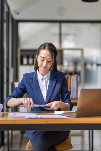 Brązowe włosy w jasnoniebieskiej kurtce uśmiechnięta Azjatka pracuje z laptopem dokumentu w biurze, robi planowanie analizy raportu finansowego, inwestycji biznes plan, koncepcja analizy finansów. - Zdjęcie, obraz