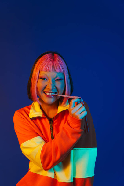 Asian girl with pink hair making fun while eating chewing marmalade isolated over blue background - Фото, изображение