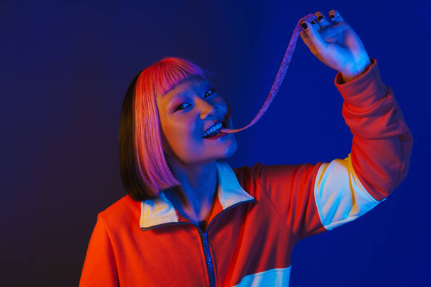 Asian girl with pink hair making fun while eating chewing marmalade isolated over blue background - Fotó, kép