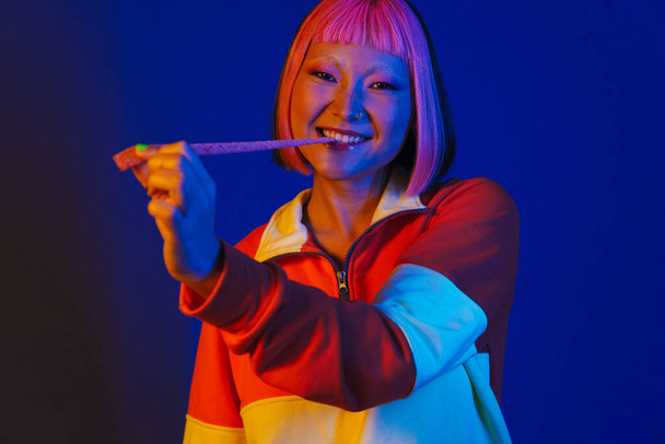Asian girl with pink hair making fun while eating chewing marmalade isolated over blue background - Fotografie, Obrázek