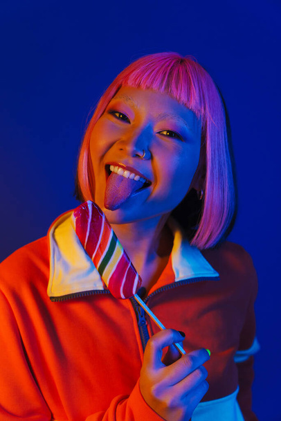 Asian girl with pink hair making fun while eating ice cream isolated over blue background - 写真・画像