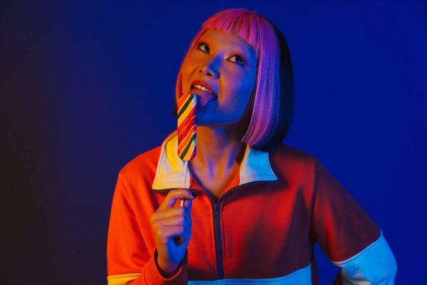 Asian girl with pink hair making fun while eating ice cream isolated over blue background - Foto, Bild
