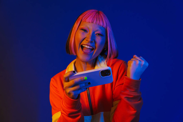 Asian girl gesturing while playing online game on mobile phone isolated over blue background - Foto, Bild
