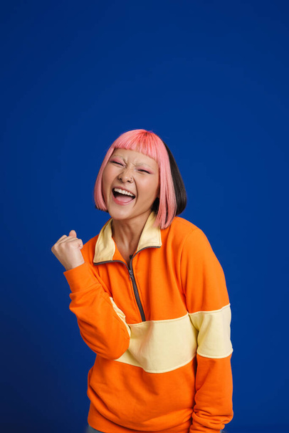 Asian girl with pink hair screaming while making winner gesture isolated over blue background - Valokuva, kuva
