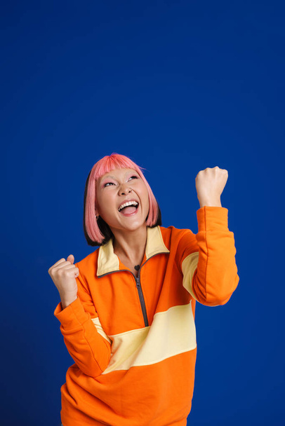 Asian girl with pink hair screaming while making winner gesture isolated over blue background - Foto, afbeelding