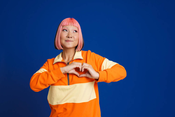 Asian girl with pink hair laughing and showing heart gesture isolated over blue background - Фото, изображение