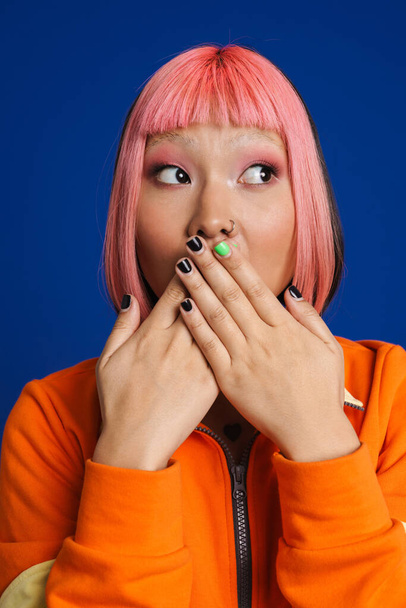 Asiatisches Mädchen drückt Überraschung aus und bedeckt ihren Mund isoliert vor blauem Hintergrund - Foto, Bild