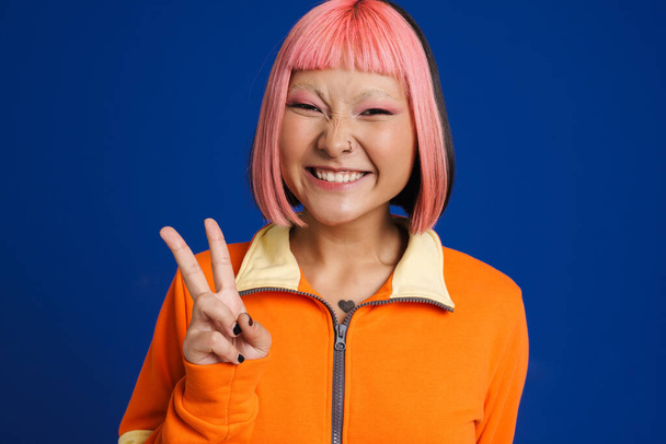 Asian girl with pink hair laughing while showing peace gesture isolated over blue background - Valokuva, kuva