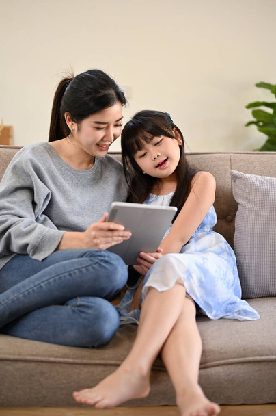 Retrato, Hermosa y amable mamá asiática está viendo videos o jugando un juego con su hija pequeña en una tableta mientras se relaja en la sala de estar juntos. - Foto, imagen
