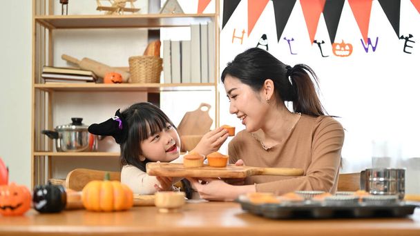 Gioiosa e giocosa bambina asiatica in costume di Halloween si sta divertendo molto con sua madre, dando da mangiare cupcake a sua madre. Concetto di tempo famiglia felice - Foto, immagini