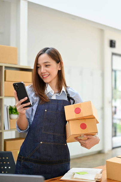 Portre, Çekici Asyalı bin yıllık bayan küçük online iş sahibi kargo kutuları tutuyor ve müşterilerden gelen bir mesajı okumak için telefonunu kullanıyor.. - Fotoğraf, Görsel