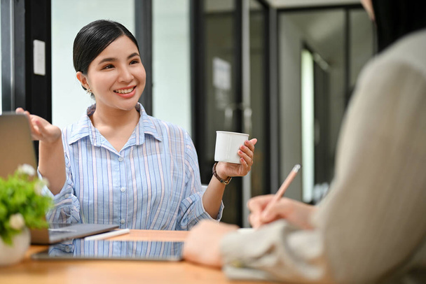Όμορφη και χαρούμενη νεαρή Ασιάτισσα επιχειρηματίας ή υπάλληλος γραφείου απολαμβάνει να μιλάει με τη συνάδελφό της κατά τη διάρκεια του διαλείμματος καφέ στο γραφείο. - Φωτογραφία, εικόνα
