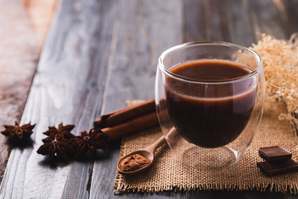 Heißer Kakao im Becherglas mit Schokolade, Zimtstange, Sternanis und Kakaopulver auf Holzgrund, Heißgetränk im Winter - Foto, Bild