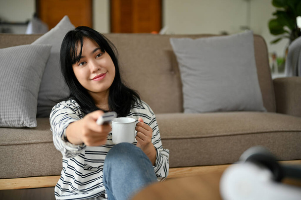 Hezká asijská žena se ráda dívá na film nebo televizní pořad v televizi, zatímco popíjí ranní kávu v obývacím pokoji. Koncept životního stylu - Fotografie, Obrázek