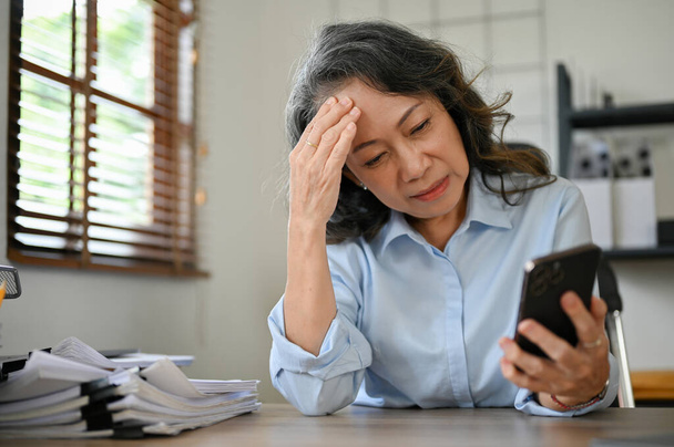 Stressed and sad Asian aged businesswoman or senior female manager at her desk, staring at her smartphone screen, receiving bad news, having problems with her supplier - Photo, Image