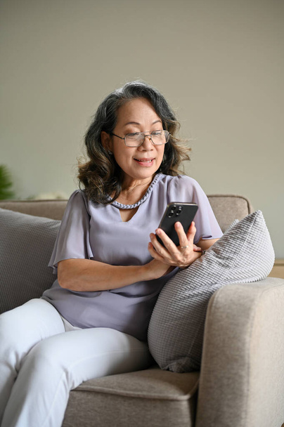 Πορτρέτο, Όμορφη ηλικιωμένη Ασιάτισσα γυναίκα φορώντας γυαλιά ηλίου, κάθεται στον άνετο καναπέ της στο σαλόνι, διαβάζοντας online ειδήσεις στο κινητό της τηλέφωνο. - Φωτογραφία, εικόνα