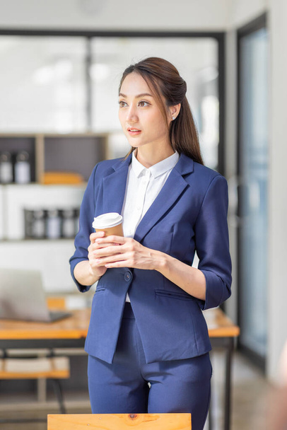 Portrait of young Asian woman hoding coffee cup and standing in a busy modern workplace - Zdjęcie, obraz