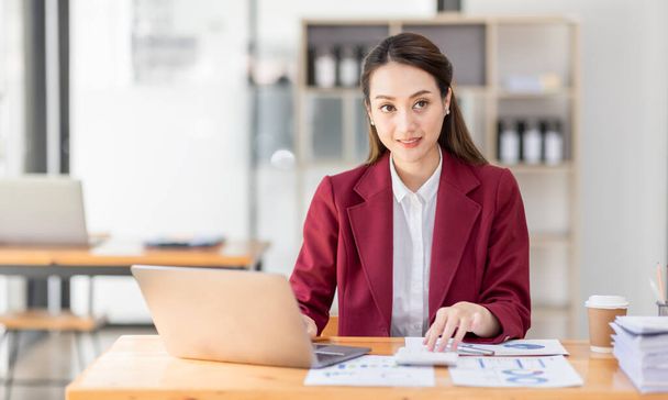 職場でラップトップを使用しているアジアの女性,赤いジャケットスーツを身に着けているビジネス女性は、お金、投資、経済、節約お金や保険の概念をチェックノートパソコン上で忙しい作業 - 写真・画像