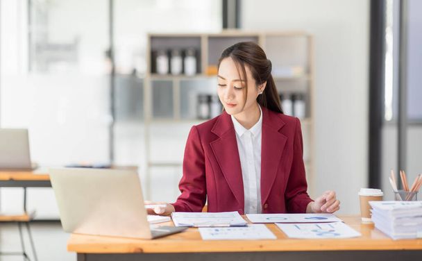 Азійка працює ноутбуком на робочому місці, жінка в костюмі червоної куртки зайнята роботою над комп'ютерними чеками Фінанси, інвестиції, економіка, заощадження грошей або страхова концепція - Фото, зображення