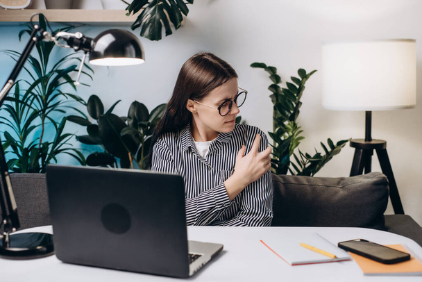 Unhappy young woman in eyeglasses with hand holding shoulder pain. Office syndrome and Health care concept. Businesswoman working with laptop feeling shoulder pain sitting on couch at home office - Foto, Bild