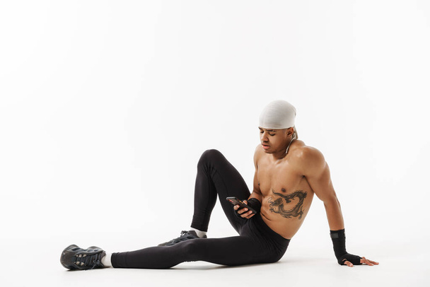 Mladý sportovec bez košile s tetováním pomocí mobilu během cvičení izolované přes bílé pozadí - Fotografie, Obrázek