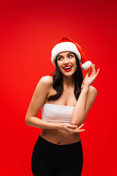 Smiling brunette woman in top and santa hat posing isolated on red - Foto, Bild