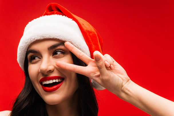 Cheerful young woman in santa hat showing victory sign isolated on red - Photo, Image