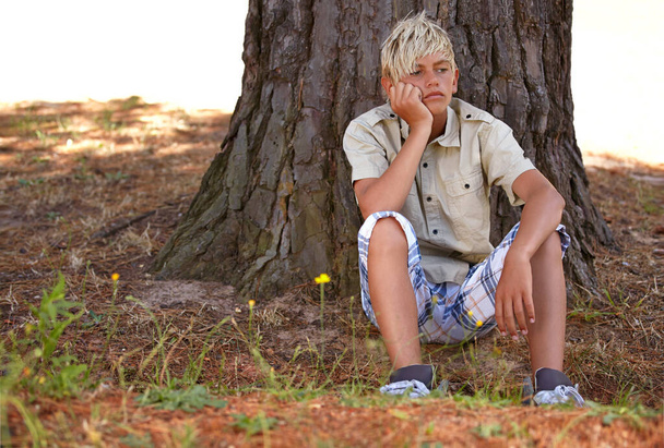 Le camp d'été craint. Un jeune garçon assis sous un arbre regardant loin avec une expression ennuyeuse - Photo, image