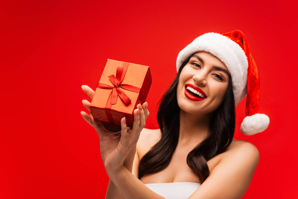 Cheerful young woman in santa hat holding gift box isolated on red - Fotografie, Obrázek