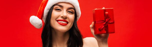 Portrait of positive woman in santa hat holding present with bow isolated on red, banner  - Photo, Image