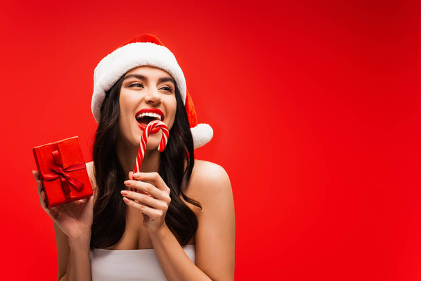 Excited woman in top and santa hat holding striped lollipop and present isolated on red - Photo, image