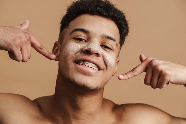Young shirtless man with cosmetic clay mask pointing fingers at his smile isolated over beige background - Zdjęcie, obraz