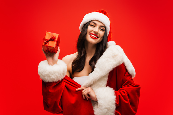 Positive model in santa costume holding present and looking at camera isolated on red - Photo, Image