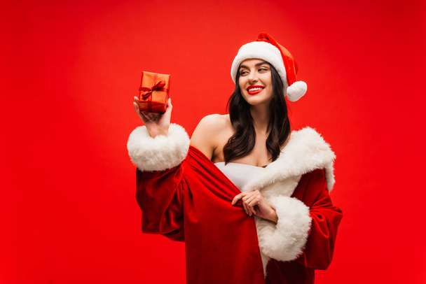 Hezká brunetka žena v kostýmu Santa při pohledu na současnost s stuhou izolované na červené - Fotografie, Obrázek