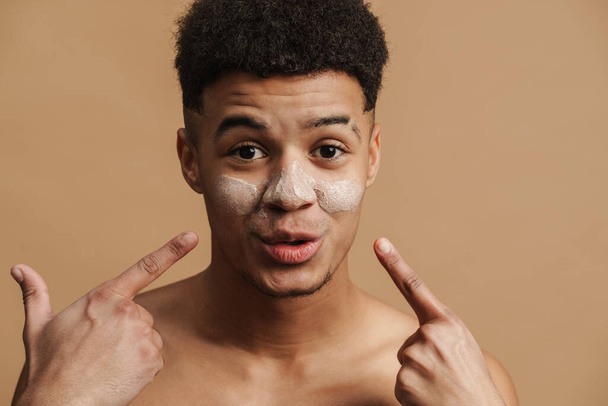 Young shirtless man with cosmetic clay mask pointing fingers at his face isolated over beige background - Фото, зображення