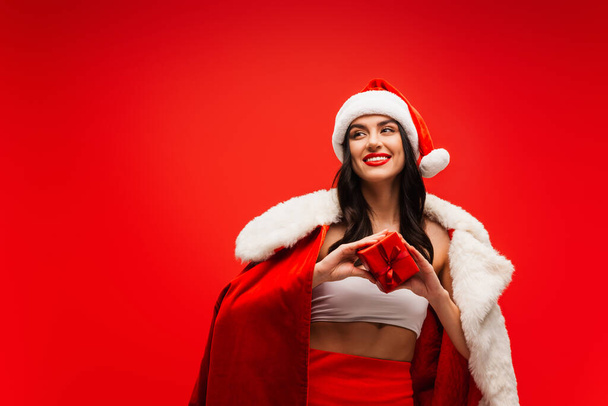 Pretty young woman in santa costume and hat holding gift box isolated on red - Zdjęcie, obraz