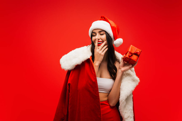 Young woman in santa costume and hat holding present and touching face isolated on red - Фото, зображення