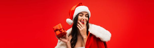 Hübsches brünettes Model in Weihnachtsmannkostüm und Hut mit Geschenk und berührendem Gesicht auf rotem Banner  - Foto, Bild