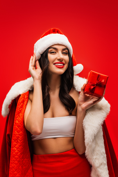 Smiling woman in santa costume holding present with bow isolated on red - Foto, imagen