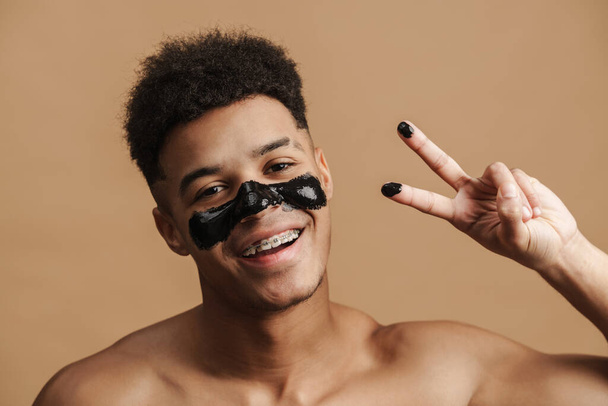 Shirtless man with cosmetic mask smiling and gesturing at camera isolated over beige background - Foto, afbeelding