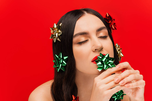 Young brunette woman with visage holding gift bows near lips isolated on red  - Fotó, kép