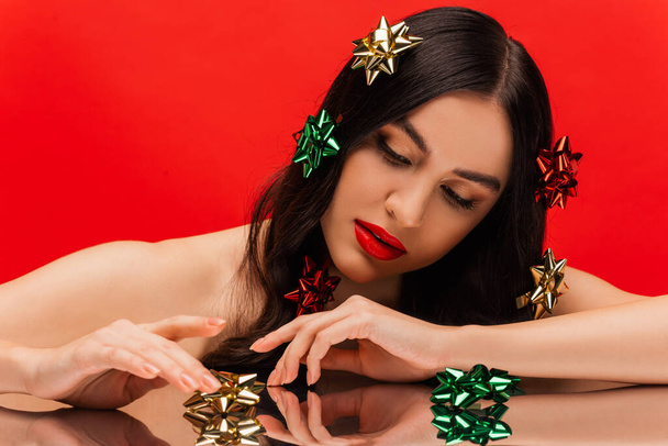 Pretty woman with naked shoulders and shiny gift bows touching reflective surface isolated on red  - Fotografie, Obrázek